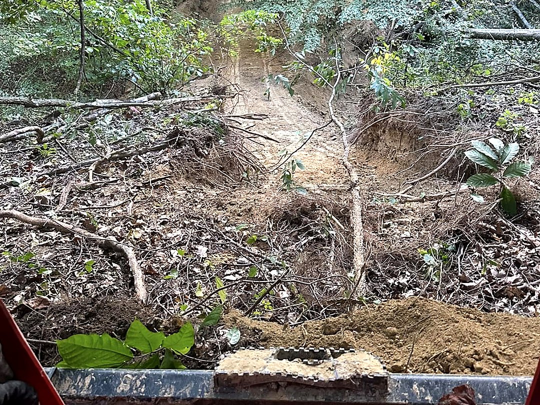 View over the bucket while cutting an access track up a wooded slope, brush and timber pushed back on both sides