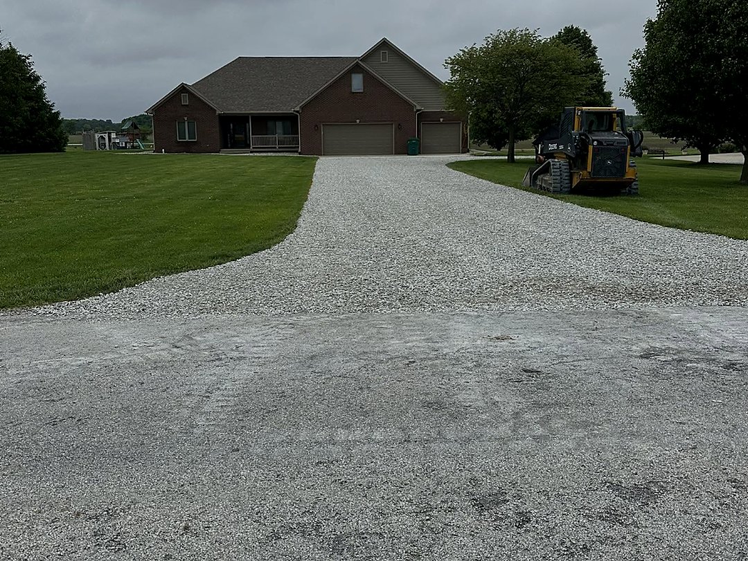 Wide gravel drive leading to a brick ranch house with a skid steer parked at the treeline