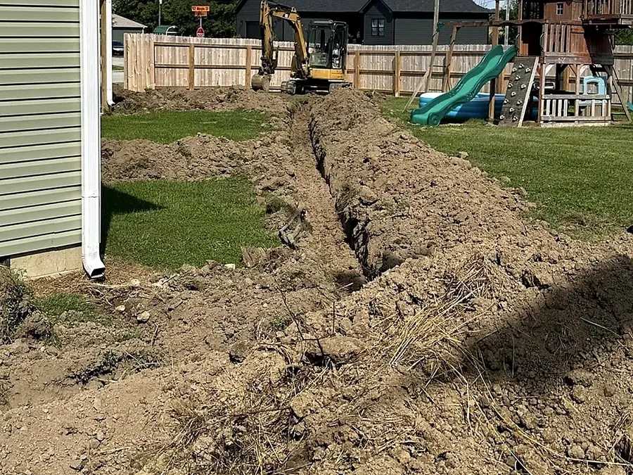 Excavator digging a drainage trench across a back yard beside a play set